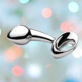 The Njoy Pure Butt Plug Large, a sleek, silver, hypoallergenic accessory featuring an anchor-shaped design with a teardrop-shaped bulbous end and a ring handle for easy removal, is showcased against a soft, multicolored background adorned with bokeh lights.